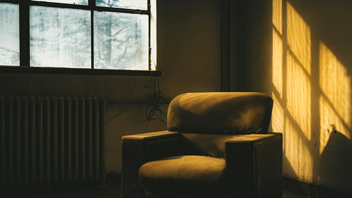 An old armchair in an empty room, lit by low golden light through a window, with the window frame casting shadows on the wall
