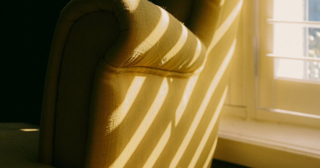 An empty pale armchair beside a window, with bands of warm late sunlight falling across it