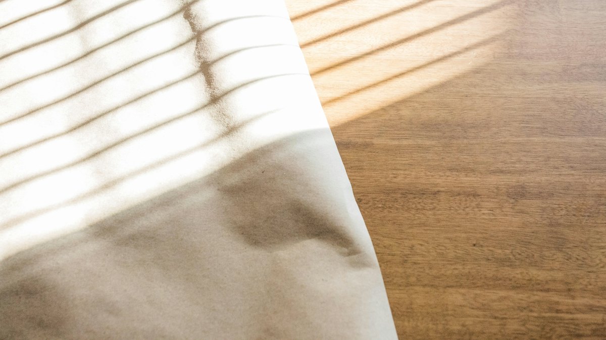 A blank sheet of pale paper lying on a warm wooden table in striped daylight