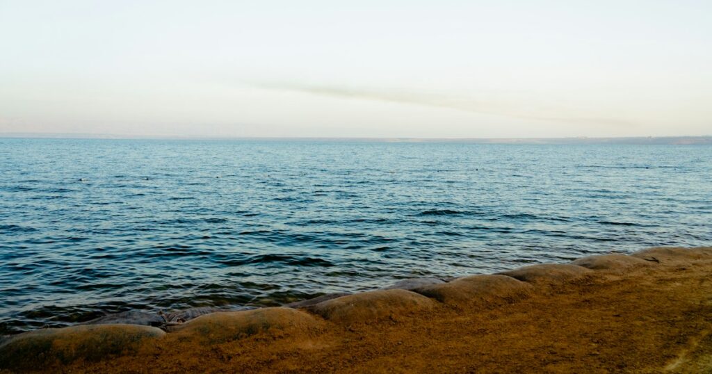 Calm open water seen from a sandy shore, with a far coastline on the horizon in pale evening light