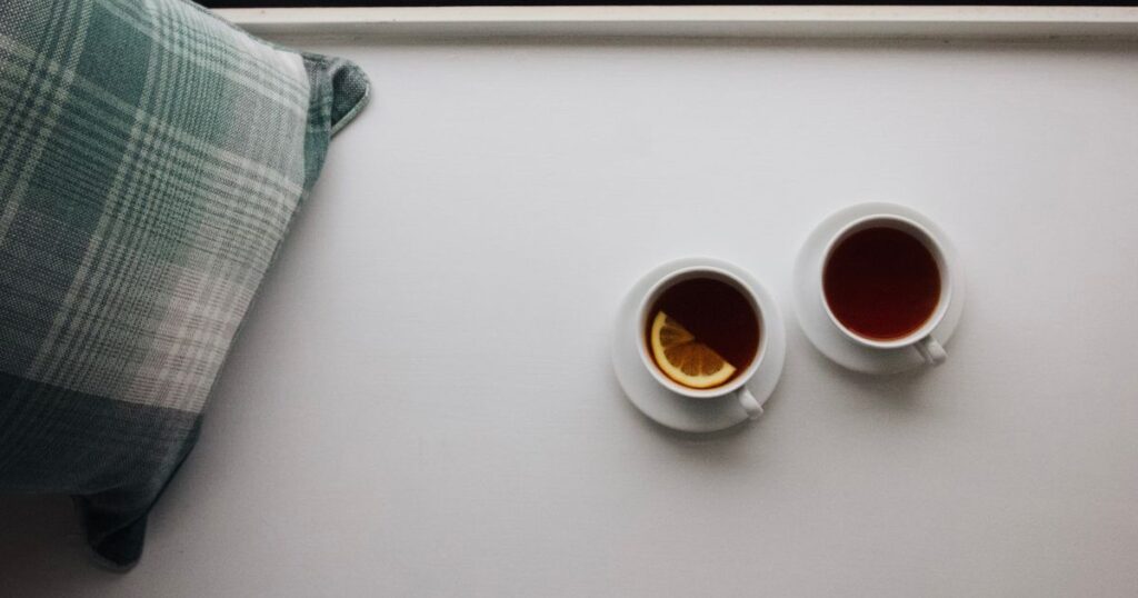 Two cups of tea side by side on a white windowsill, one with a slice of lemon, next to a green checked cushion