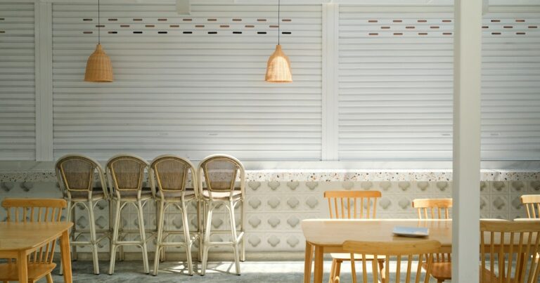 Empty wooden chairs and stools lined up along a pale panelled wall in daylight