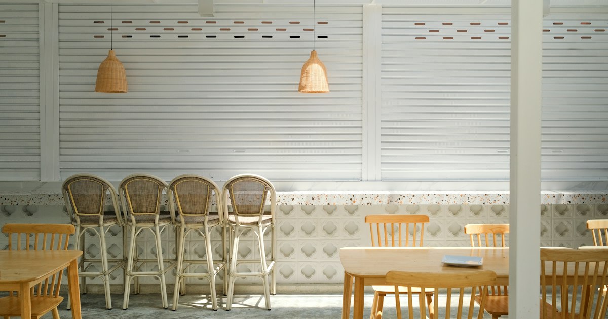 Empty wooden chairs and stools lined up along a pale panelled wall in daylight