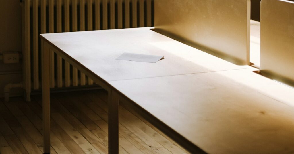 A long pale desk in an empty room, lit by low afternoon sun, with a single sheet of paper lying on it and a wooden floor below
