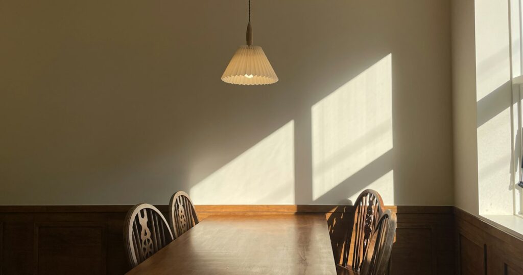 A long wooden dining table with chairs on both sides in an empty room, lit by daylight from a tall window