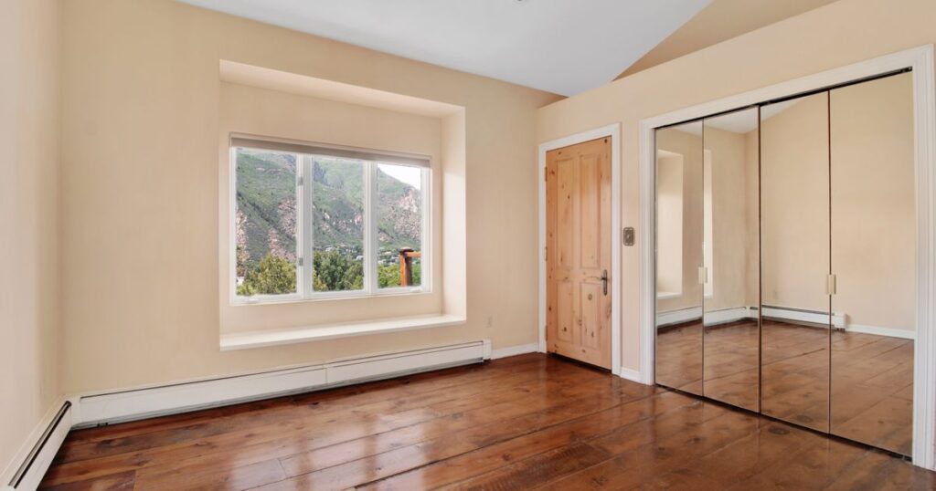 An empty room with a polished wooden floor, a wide window looking out to green hills, and a pale door standing closed