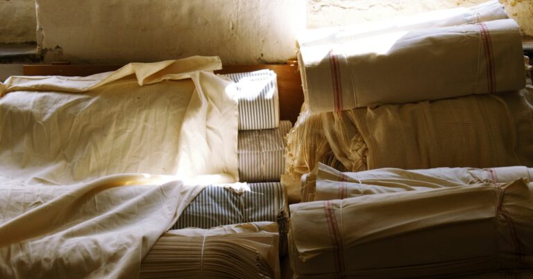 Stacks of folded and tied linen cloth against a whitewashed wall, lit by a shaft of daylight