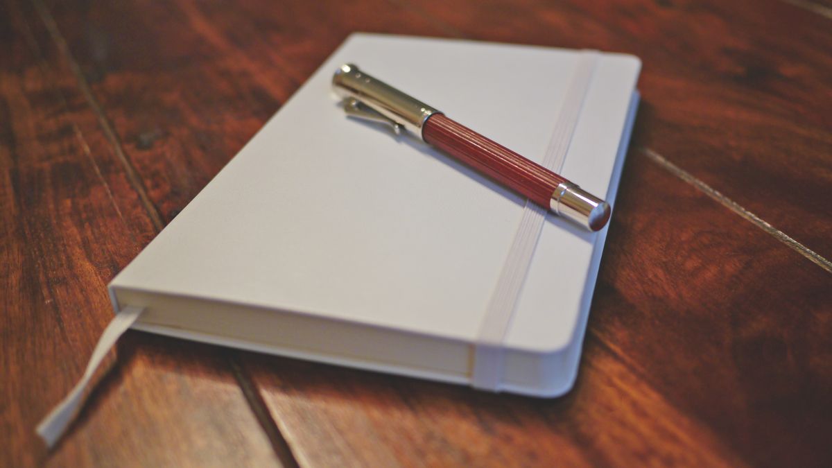 A closed white notebook with a pen resting on it, on a wooden table