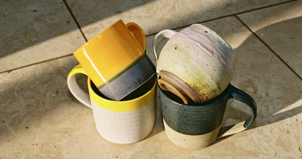 Four handmade stoneware mugs in different glazes resting together in low golden sunlight on a stone floor