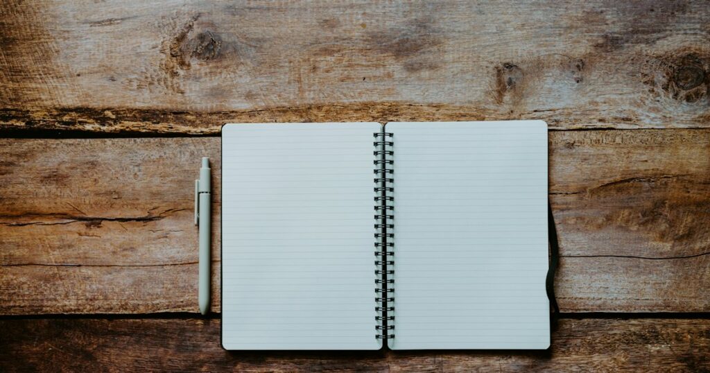 An open blank spiral notebook and a pen lying on a worn wooden table