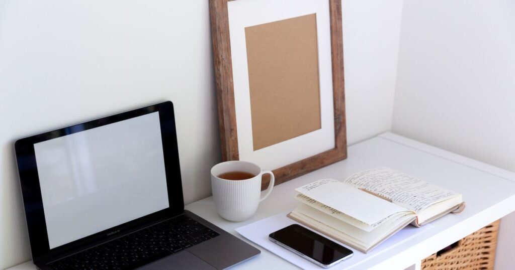 An open laptop with a blank screen beside a cup of tea, a phone and an open notebook on a white desk, with an empty picture frame against the wall