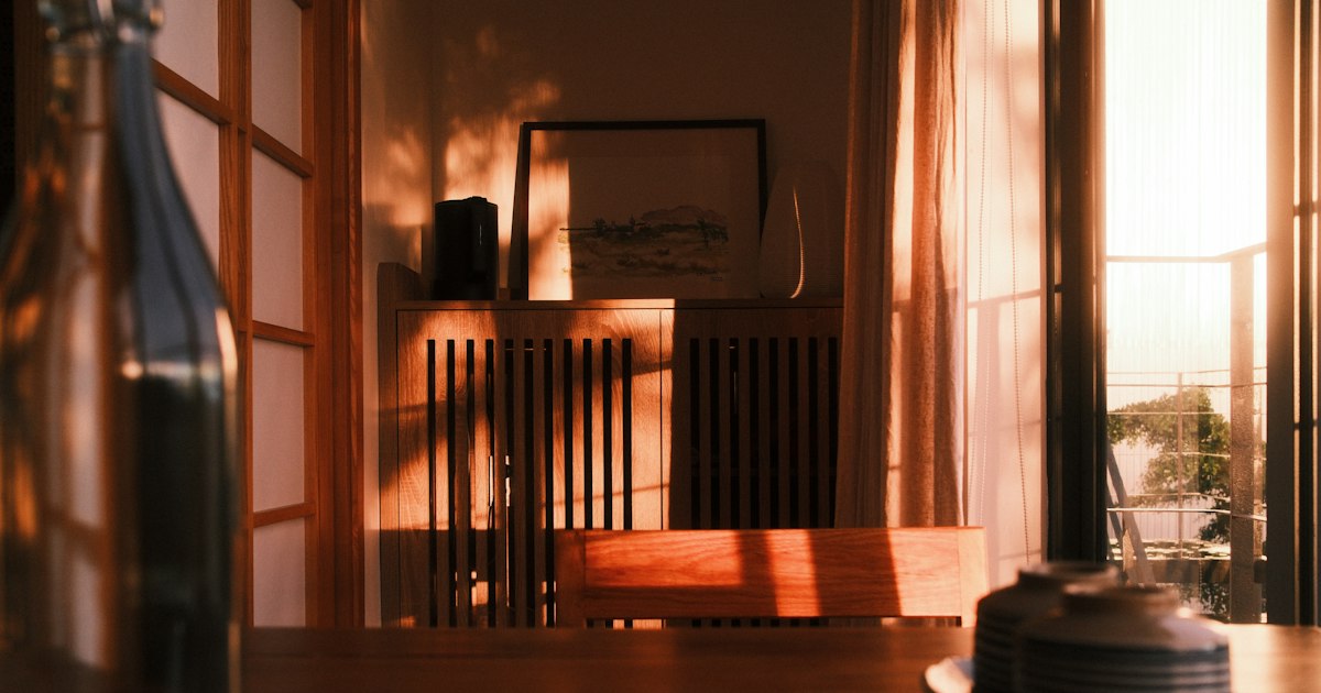 Late afternoon light falling across a wooden dining room, with an empty chair and a curtained window