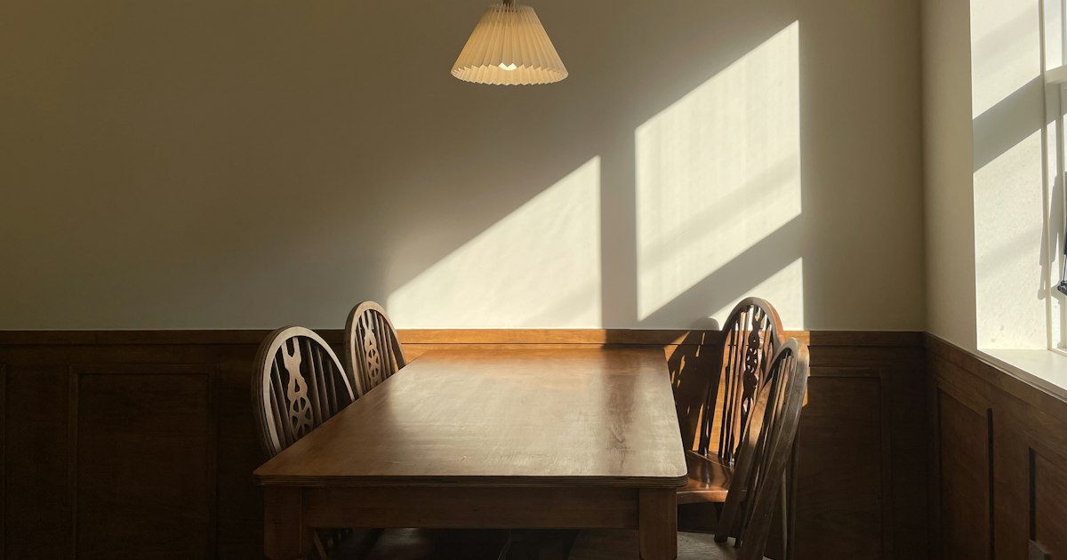 An empty wooden dining table with chairs on both sides, lit by low sun through a window