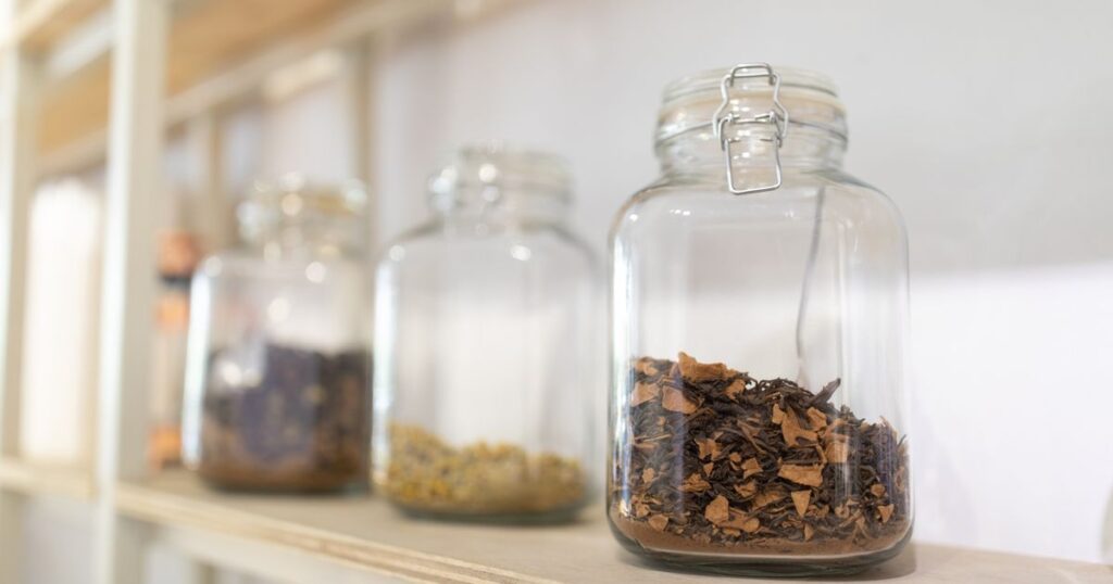 Glass storage jars of dried herbs lined up on a pale wooden kitchen shelf
