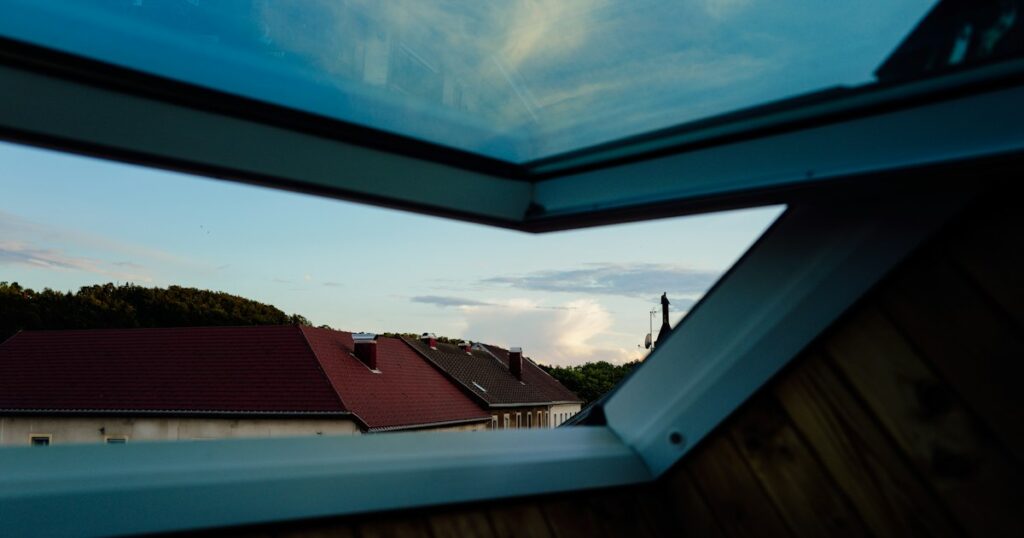 An open attic window looking out over tiled rooftops towards trees and an evening sky