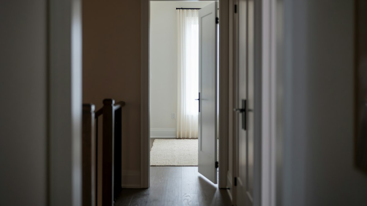 A hallway with an open door leading into a bright room with a pale curtain at the window