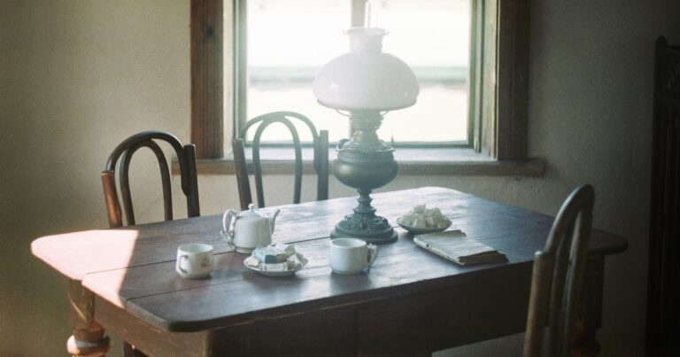 Pale daylight on a wooden table laid with cups and a lamp, the chairs around it empty