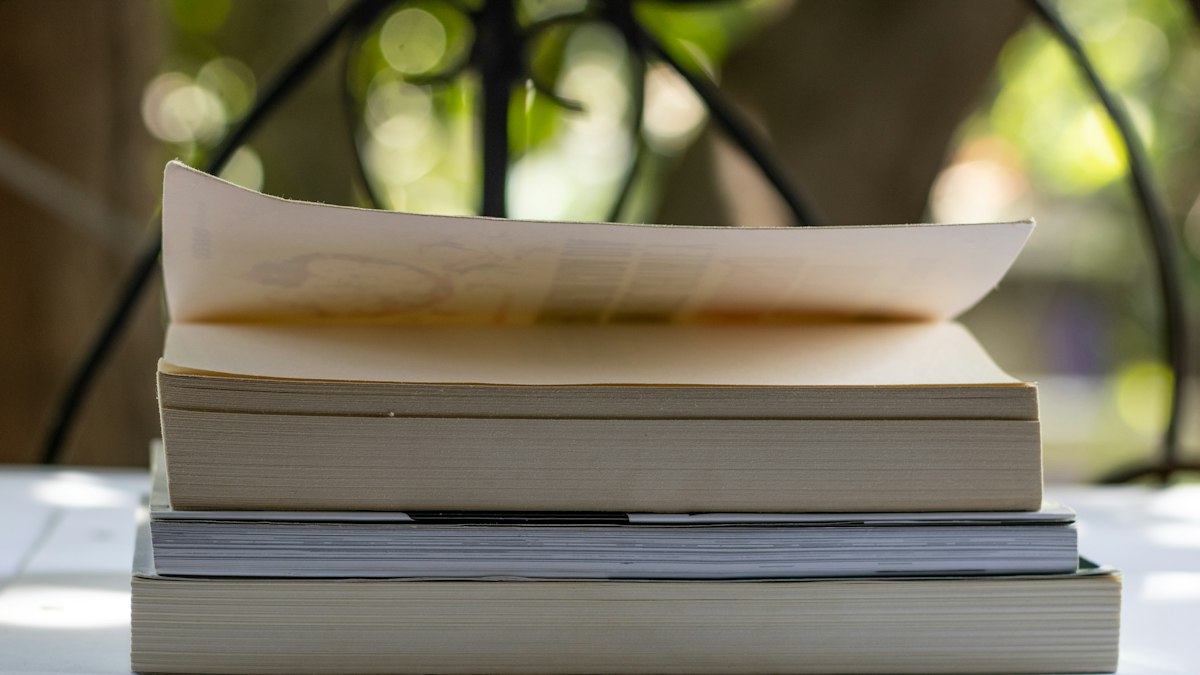 A small stack of paperback books on a pale table outdoors, the top one open and curling, with soft green foliage blurred behind