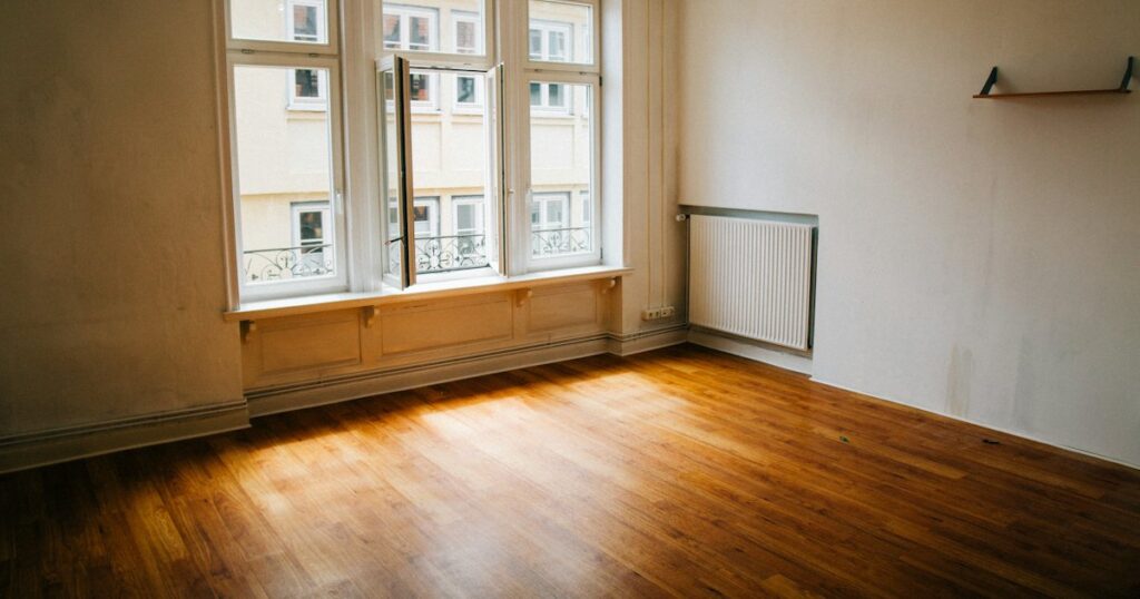 An empty room with cream walls and a warm wooden floor, daylight falling through a tall window