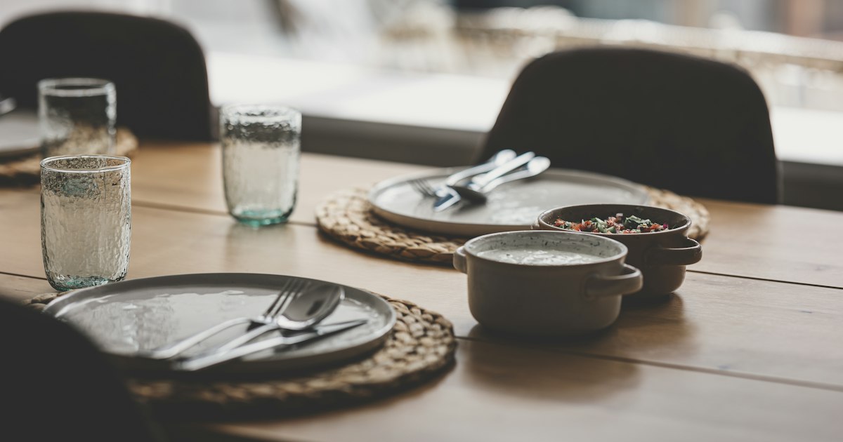 A wooden table laid with plates, glasses and two serving bowls, chairs pulled up to it, daylight from a window behind