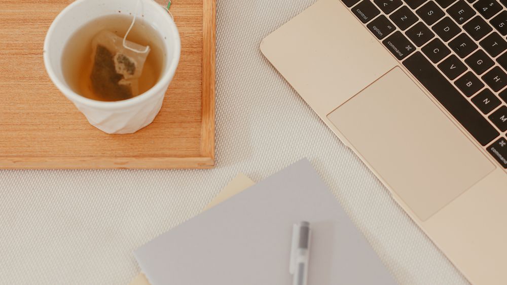 A cup of tea on a wooden tray next to an open laptop and a grey notebook with a pen, seen from above