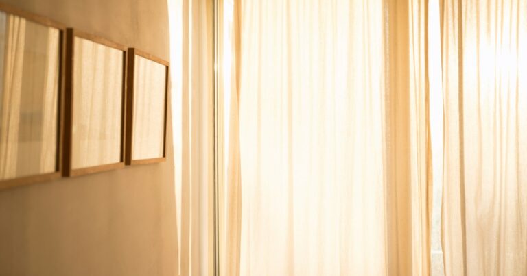 Three empty wooden frames on a pale wall beside a sunlit curtain