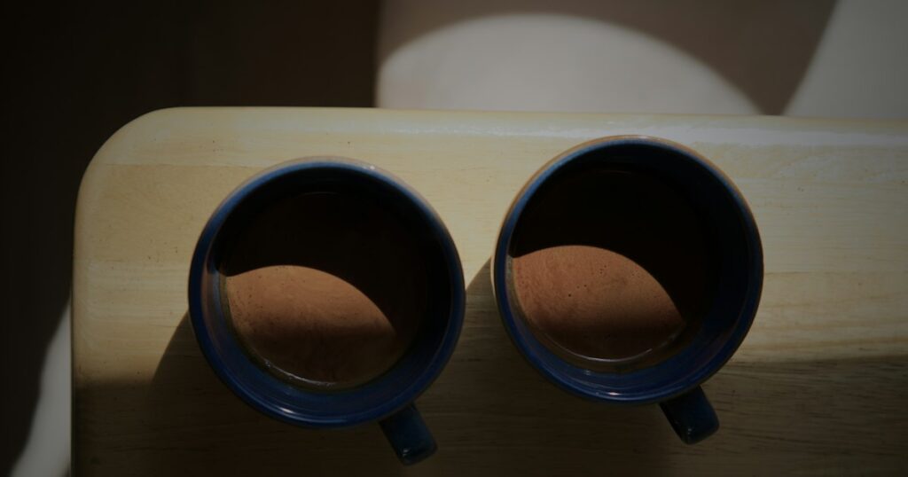 Two blue stoneware cups of coffee side by side on a pale wooden board in a patch of sunlight