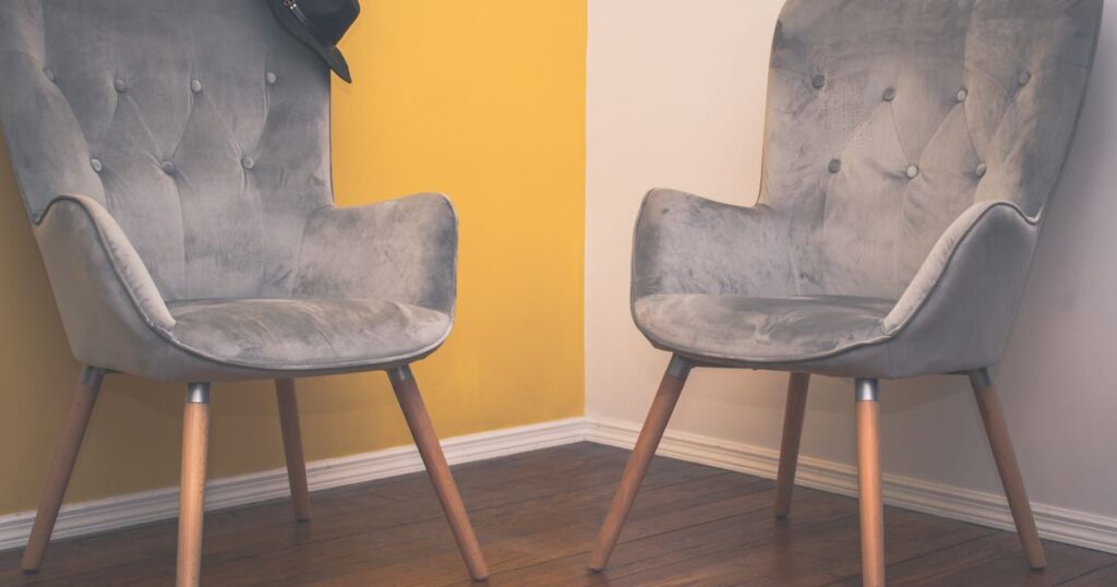 Two grey upholstered armchairs angled towards each other on a wooden floor, against a white and mustard yellow wall