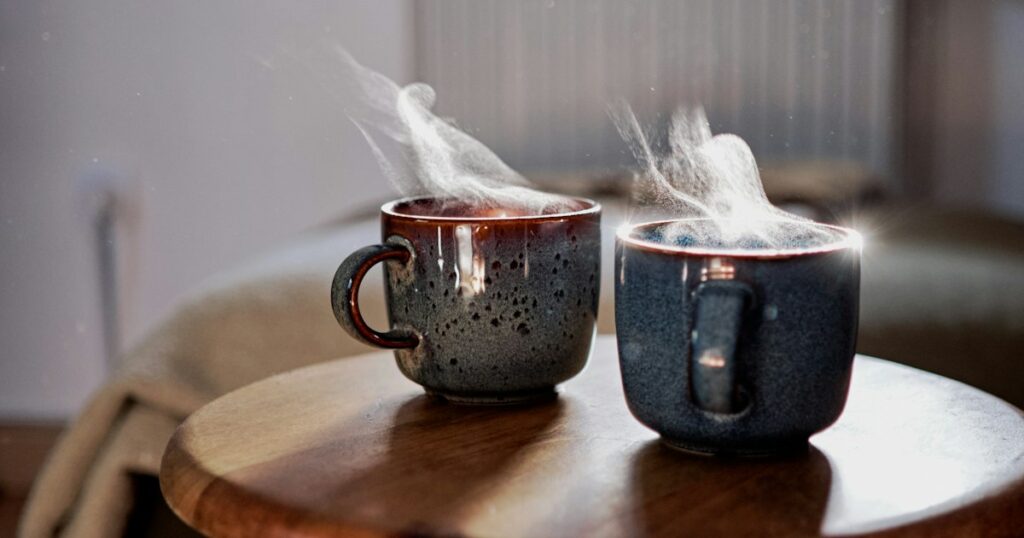Two steaming ceramic mugs on a small round wooden table in a quiet sunlit room