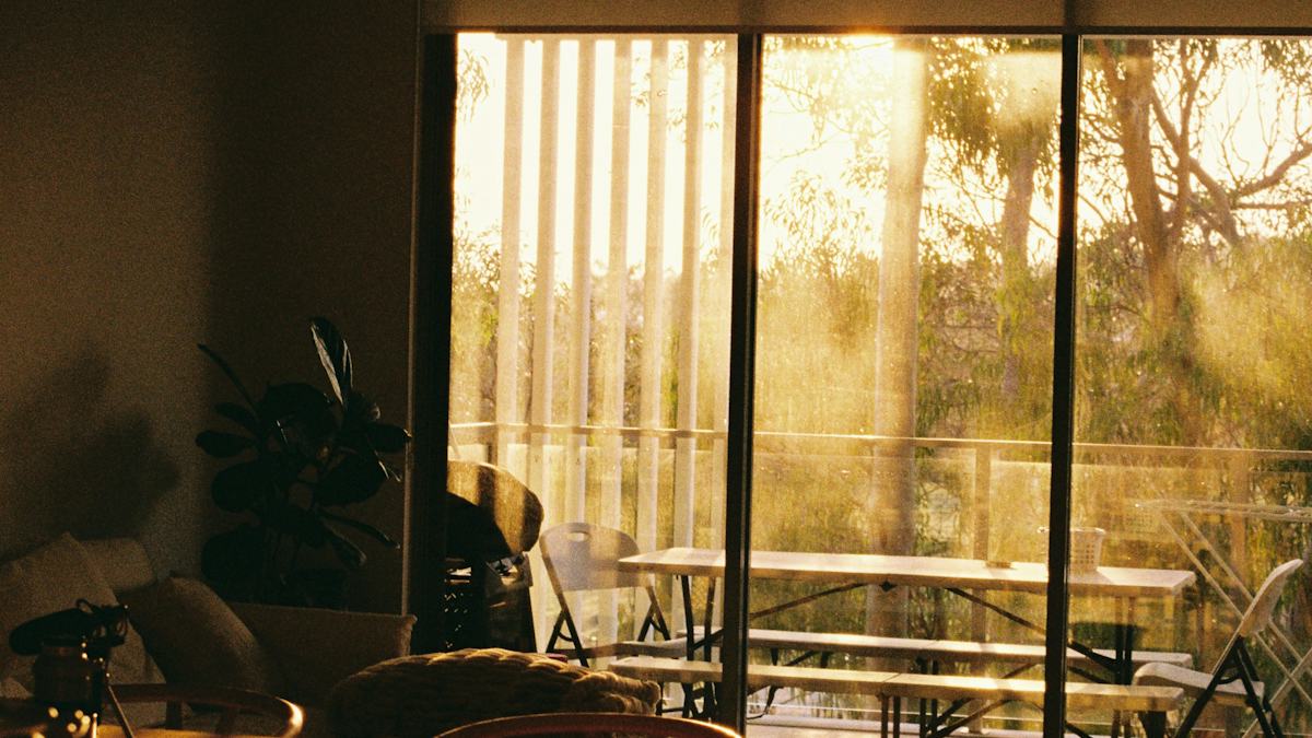 A warm living room at sunset, looking through glass doors to an empty table and benches on a balcony