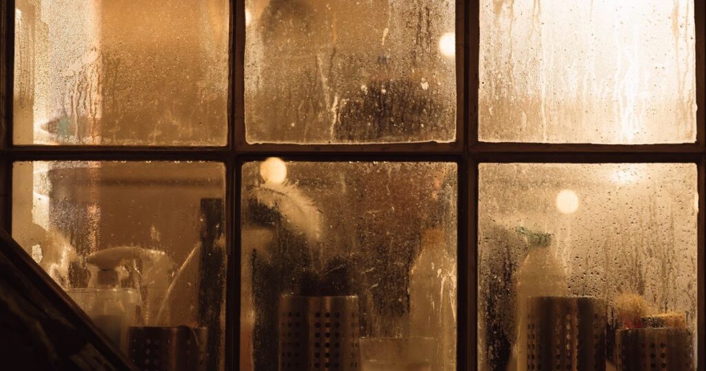 A fogged window pane with warm light behind it, the room beyond indistinct