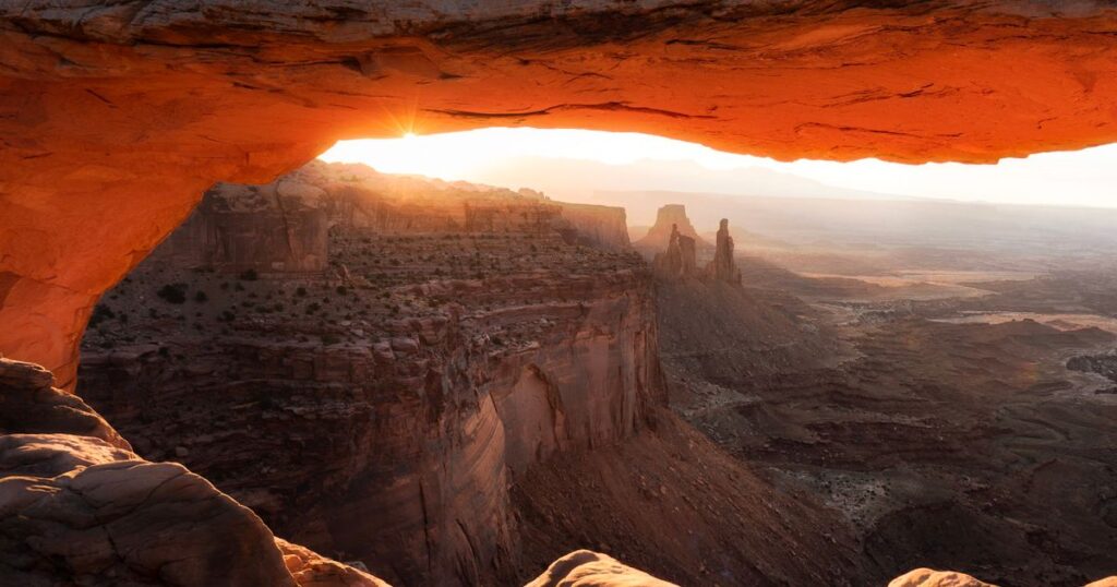 Sunrise seen from under a wide sandstone arch glowing orange above a canyon
