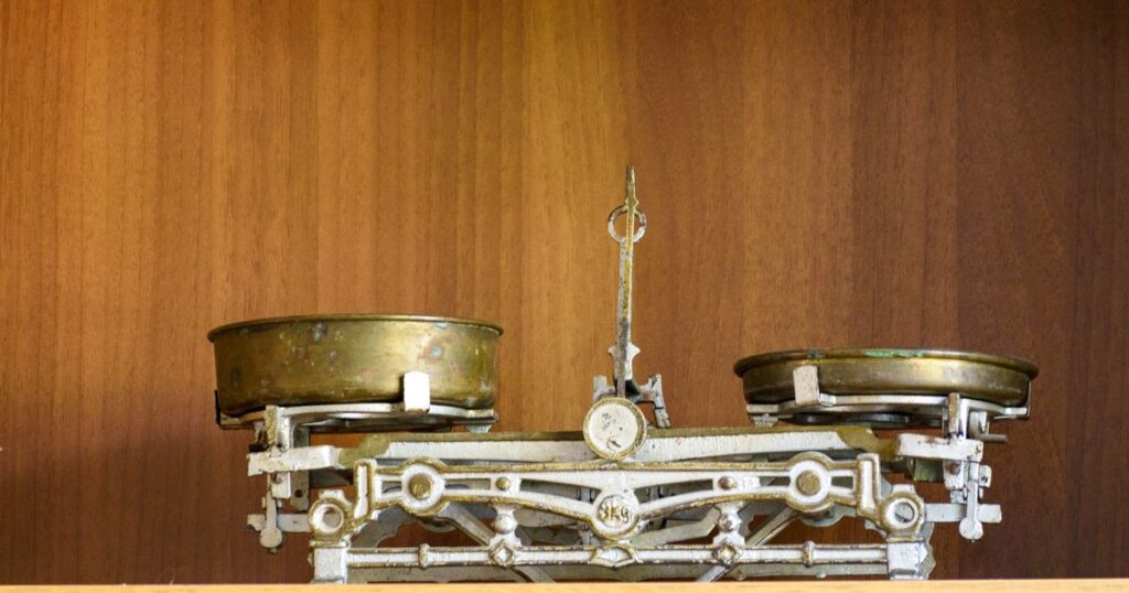 A vintage brass balance scale standing on a wooden shelf with both pans empty.