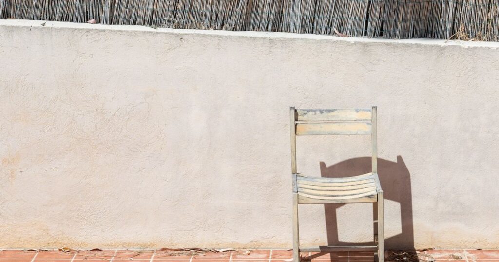 A single wooden chair against a warm pale wall, casting a sharp shadow